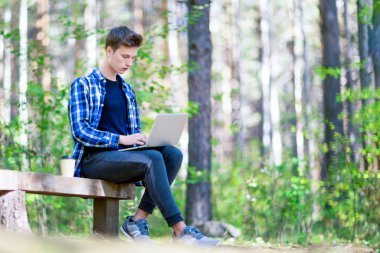 Bir yeşil orman parkta bir dizüstü bilgisayar üzerinde çalışan bir genç çocuk.