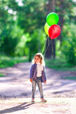 Küçük bir kız ellerinde balonlar arka planı yeşil ağaçlar orman içinde tutar..