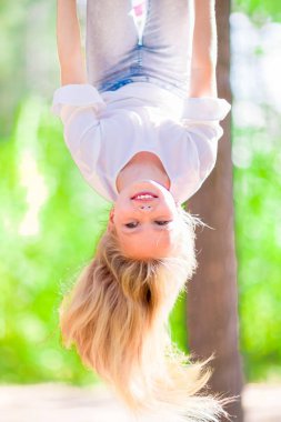 Uzun sarı saçlı neşeli genç kız baş aşağı parkta açık havada asılı. Güneşli yaz gün.