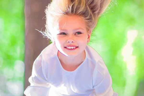 Cheerful young girl with long blond hair hangs upside down outdoors in the park. Sunny summer day.