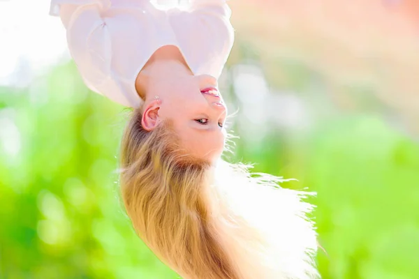 Cheerful young girl with long blond hair hangs upside down outdoors in the park. Sunny summer day.