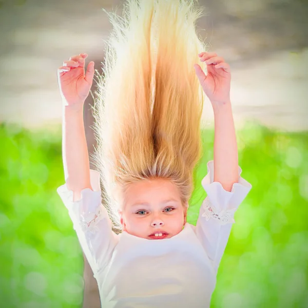 Cheerful young girl with long blond hair hangs upside down outdoors in the park. Sunny summer day.