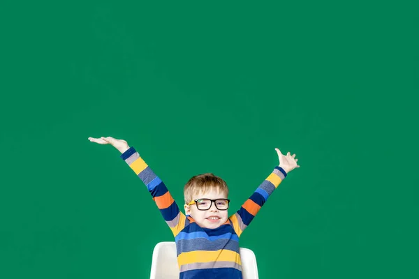 Усміхнений хлопчик в окулярах сидить за столом, підняв руки — стокове фото