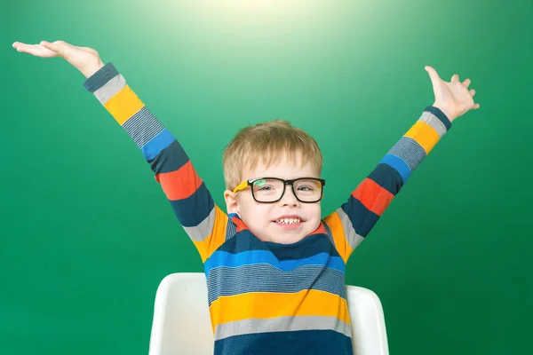 Smiling boy in glasses is sitting at the desk, raised his hands — Stock Photo, Image
