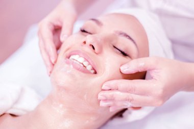 Kozmetoloji. Maske ile yüz masajı yaparken Kozmetolog elleri yakın. Spa prosedürinde gülümseyen kız