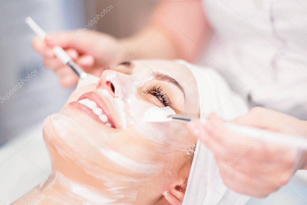 Cosmetology. The cosmetologist applies a cleansing face mask. Smiling girl on the procedure for facial rejuvenation. Spa procedure