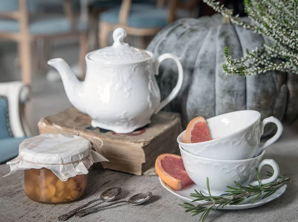Kettle, jam, old book and wo tea cups with rosemary and grapefruit on the background of a large pumpkin and heather. Tea. Autumn.
