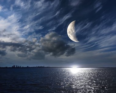 Mavi gece gökyüzünde gizemli ay, pofuduk, dramatik bulutlu gökyüzü, su dalgası üzerinde ay ışığı yansıması, ufuktaki şehir silueti, doğanın fantazisi.