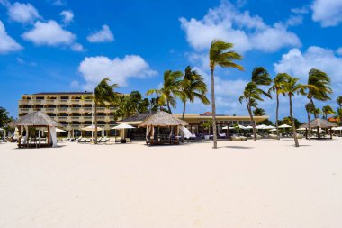 Palmiye ağaçları, beyaz plaj şemsiyeleri ve güneşli bir günde birkaç bulutla Aruba 'nın doğu yakasındaki Eagle Beach' in romantik Bucuti & Tara Resort 'unun nefes kesici manzarası.