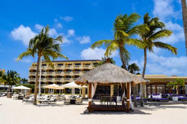 Palmiye ağaçları, beyaz plaj şemsiyeleri ve güneşli bir günde birkaç bulutla Aruba 'nın doğu yakasındaki Eagle Beach' in romantik Bucuti & Tara Resort 'unun nefes kesici manzarası.