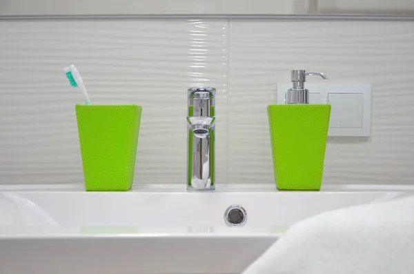 stock image Frontal view of a white sink with a shiny silver faucet, a green glass with toothbrushes and a soap dispenser and a towel.