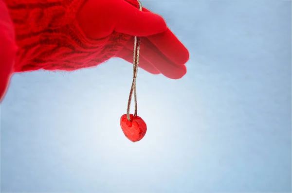 Hand in a red glove and a glass heart on a string against a blue background of snow. Image
