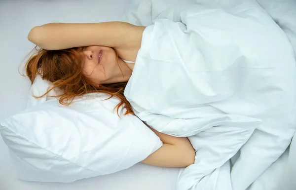 Portrait of an attractive, content, young, red-haired woman lying relaxed in bed, enjoying and snuggling with fresh, soft, white sheets and pillows in the bedroom.