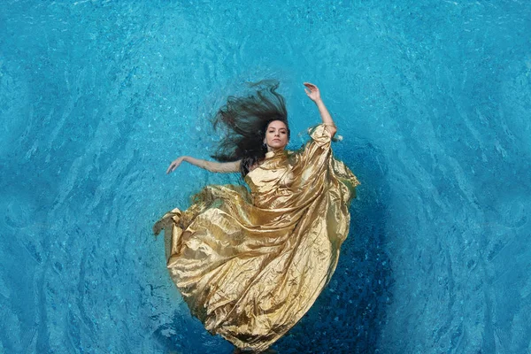 beautiful young woman in gold dress, evening dress floating weightlessly elegant floating in the water in the pool dark brown hair drift fan-shaped in turquoise water