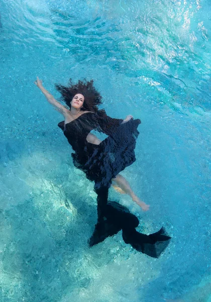 beautiful young woman in black dress, evening dress floating weightlessly elegant floating in the water in the pool dark brown curly hair floating fan-shaped in turquoise water