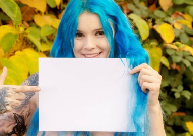 Beautiful young bohemian woman with blue hair happily holding a blank blank paper in hand, and pointing finger to it, copy space, space for your text. clipart