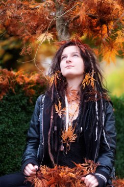 Siyah deri ceketli güzel genç bireysel, eksantrik ve kadın ile dreadlocks, piercing dövme, Japonca akçaağaç ağacın altında oturuyor ve renkli yaprakları ile mutluluk duyar