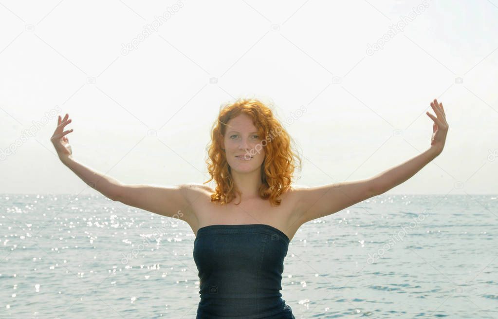 Happy happy portrait of a young elegant red-haired curly woman with outstretched arms by the sea at the beach in Liguria Italy with copy space, space for text