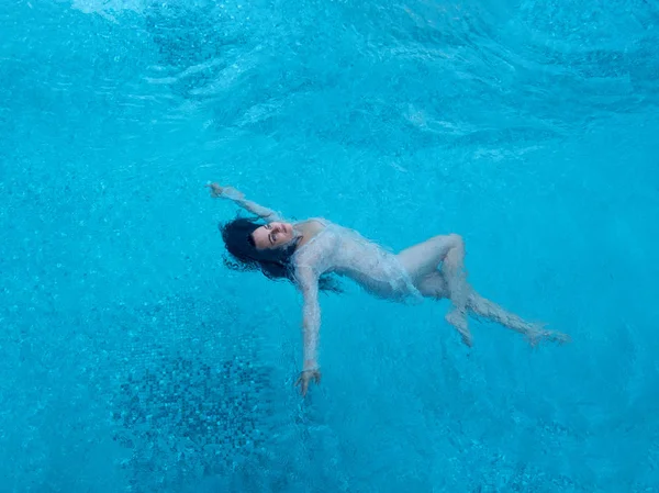 Hermosa mujer adulta en la mejor edad con pelo rizado oscuro en vestido blanco flota con elegancia flotando felizmente en agua azul turquesa en la piscina —  Fotos de Stock