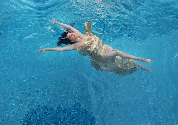 Mooie donkerharige vrouw met krullen ontspannen in gouden handdoek zweeft die gelukkig zweven in het turquoise blauwe water in het zwembad — Stockfoto