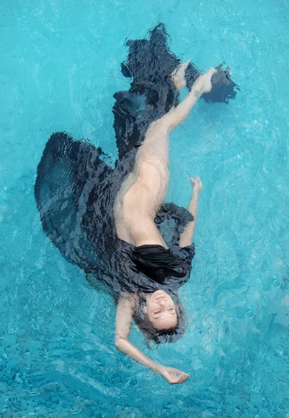 Vista superior de una hermosa joven sexy mujer de pelo oscuro relajado en vestido negro, capa, toalla flota al revés felizmente flotando en agua azul en la piscina —  Fotos de Stock