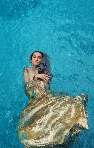 Bella giovane donna orgogliosa in abito d'oro, abito da sera galleggiante senza peso elegante galleggiante in acqua in piscina capelli castano scuro alla deriva in acqua — Foto Stock