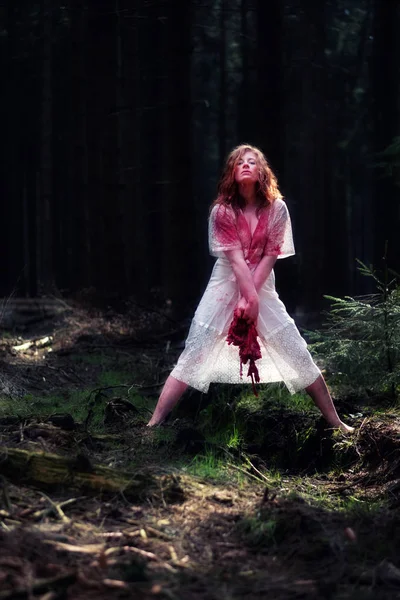 Young woman is crazy, insane in a transparent white dress, legs apart in the forest and holds a blood-smeared raw piece of meat