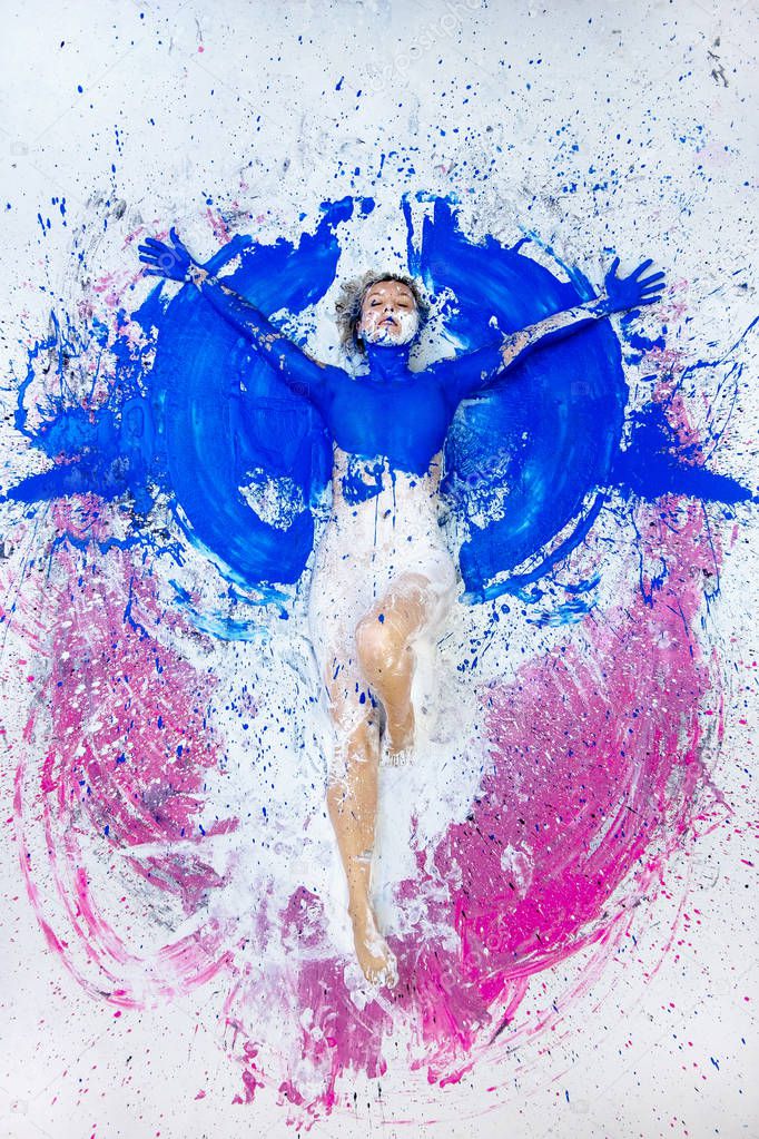 Young nude artist woman in blue, pink white, paint, painted, lies on the ground like an angel and raises her arms. Abstract body art, abstract expressionism painting,