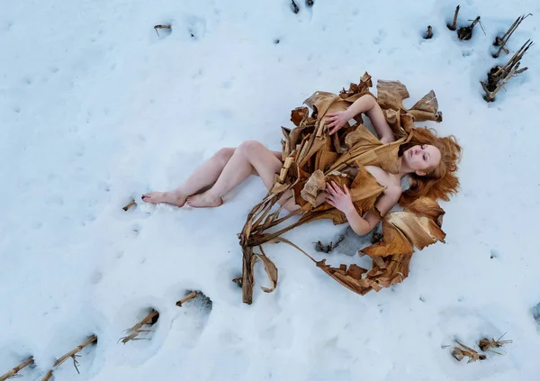 Młoda seksowna kobieta z rudymi włosami, pokryta wysuszonym drzewem bananowym leżącym na śniegu. — Zdjęcie stockowe