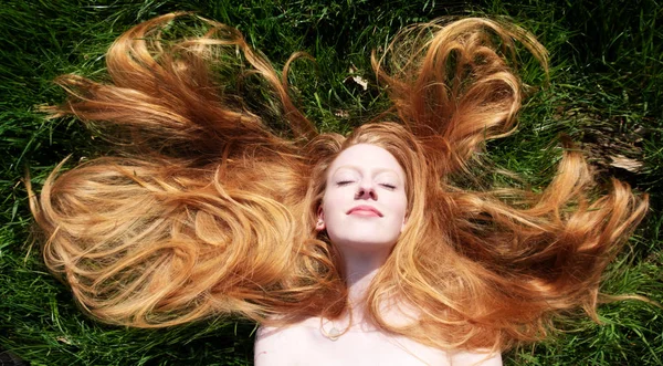 Retrato de uma bela jovem mulher sexy ruiva, deitada ao sol da primavera, relaxando na grama verde, o cabelo vermelho drapeado livremente em torno da cabeça . — Fotografia de Stock