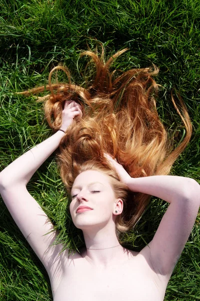 Güzel bir genç seksi kızıl saçlı kadının portresi, kollarını yükselterek, yeşil çim üzerinde rahatlatıcı bahar güneşinde yalan — Stok fotoğraf