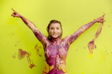 Portrait and upper body of a smiling young artistically abstract woman in pink and yellow painted against light green wall background, happily stretching both arms upwards. clipart