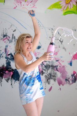 Young woman artist opens a sparkling bottle of champagne in color-stained white undershirt and is surprised