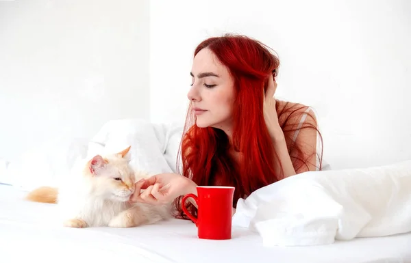Attractive, contented, young, sexy red-haired woman lying relaxed in bed with her cat with her coffee in a red cup, copy space.