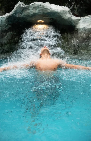 Jonge man geniet van het vallende water onder de waterval in het zwembad — Stockfoto