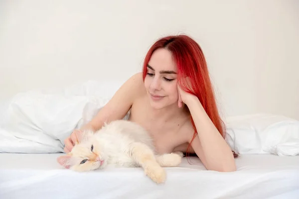 Portrait of an attractive, contented, young, sexy, red-haired woman lying relaxed in bed, cuddling her cat, copy space