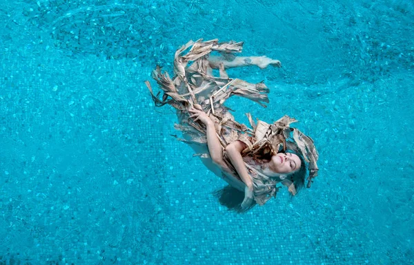 Vue du dessus d'une belle jeune femme sexy aux cheveux foncés détendue dans des feuilles desséchées, enveloppée dans un bananier, flotte joyeusement flottant dans l'eau turquoise bleue dans la piscine — Photo