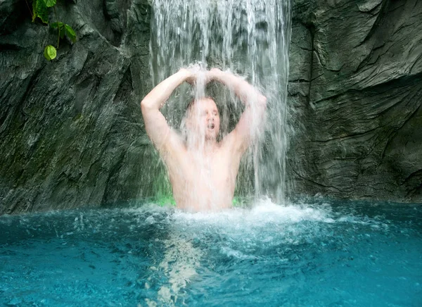 Jonge man geniet van het vallende water onder de spetterende waterval in het turquoise spabad — Stockfoto