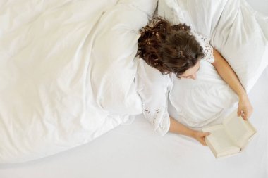 Top View çekici, memnun, genç, seksi koyu saçlı kadın yatakta rahat yatarken, zevk ve taze, yumuşak, beyaz çarşaf ve yastıkbir kitap okuma