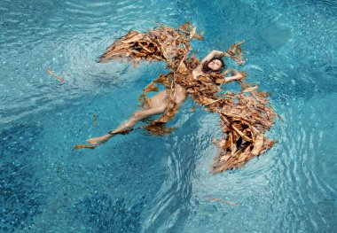 Top view of a beautiful sexy woman floating relaxed in dried up leaves of a banana tree, in shape of wings as an angel in turquoise water in the pool, copy space clipart