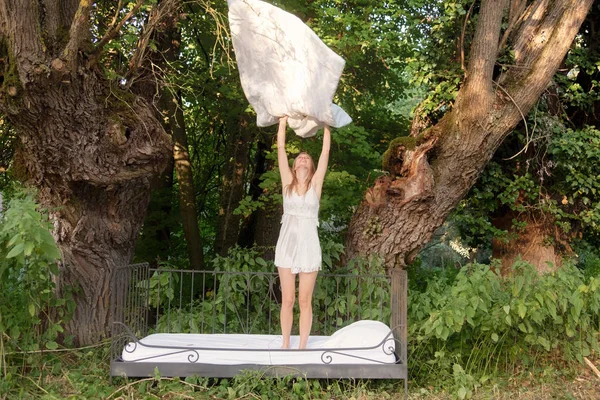 Bela jovem mulher está de pé na cama na natureza e sacudindo seu cobertor, jogando os lençóis no ar . — Fotografia de Stock