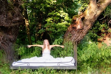 Çekici, mutlu, seksi sarışın kadın beyaz yorganlı yatakta, doğada iki yaşlı söğüt ağacının altında oturuyor.