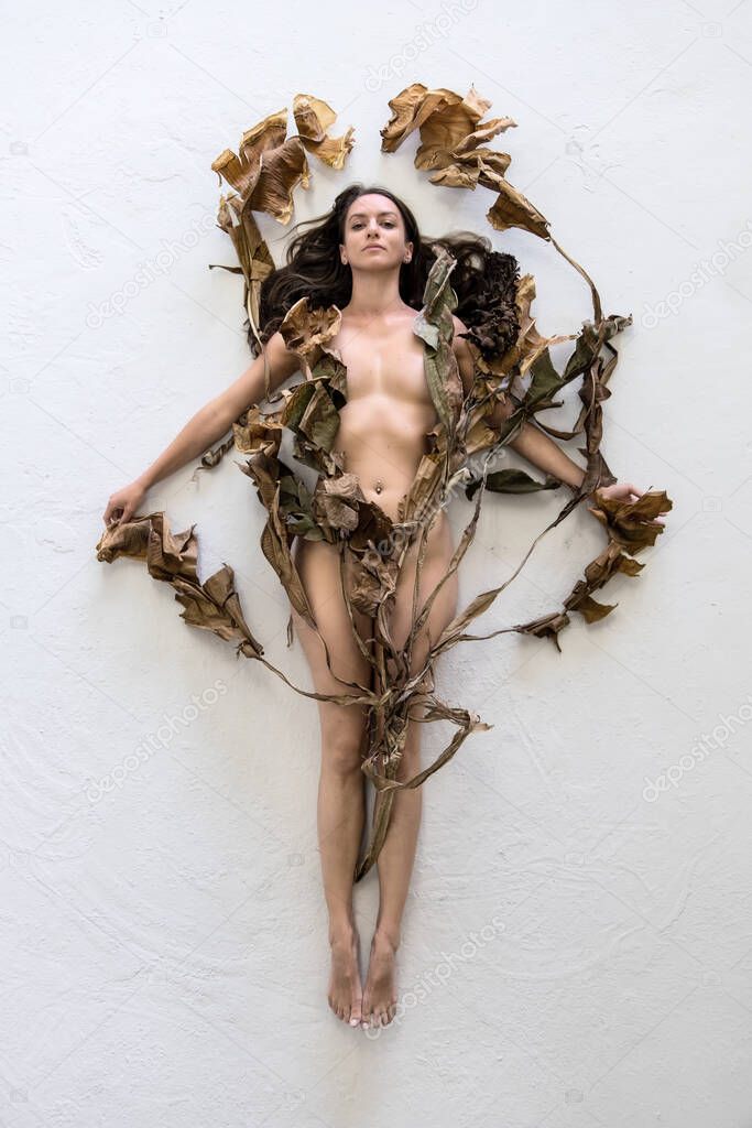 Young sexy woman with dark brown hair artfully covered with dry, withered decorative banana tree leaves lying on the gray studio floor.