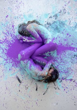 Two artistically abstract painted young sexy women, girlfriends in pink, purple, turquoise and white color lying like a Yin Yang symbol on the floor in the artists studio, copy space.