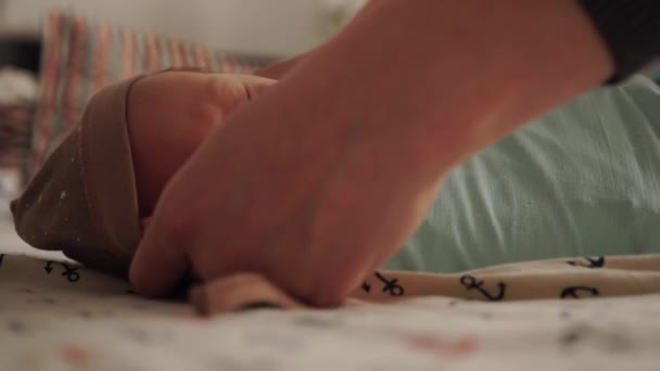 Мать пеленает своего новорожденного ребенка. Заворачивает ребенка в подгузник. Новорожденный ребенок лежит в постели. Мама надевает шляпу на ребенка — стоковое видео