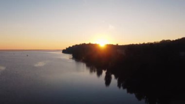 Gölün ya da nehrin üzerinde güzel bir gün doğumu. Güneş tepenin arkasından doğar. Kozalaklı ve yaprak döken ağaçlarla kıyı siluetleri. Sudaki dalgalanmalar. Güneşin doğuşu