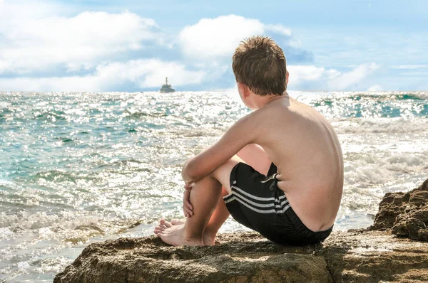 Un garçon assis sur les rochers au bord de la mer regarde le bateau naviguer — Photo