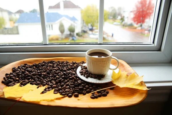 a cup of coffee with grains of coffee on a wooden plate on the windowsill opposite the window