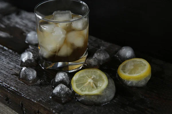Whiskey with ice in a glass on a black wooden old oak background with ice and lemon