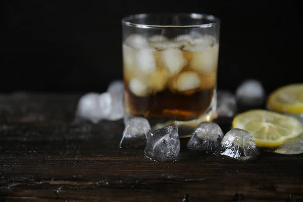 Whiskey with ice in a glass on a black wooden old oak background with ice and lemon